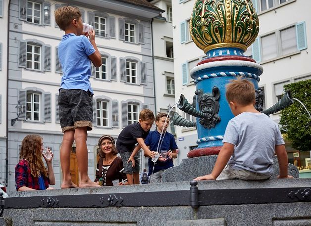 Visita guiada para crianças em Lucerna com crianças brincando e uma fonte fascinante ao fundo.