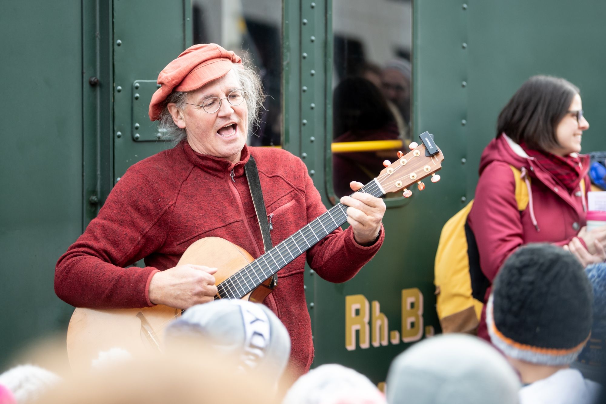 Linard Bardill gra na gitarze podczas podróży pociągiem do Krainy Kolorów, radosne osoby, kolorowe otoczenie.