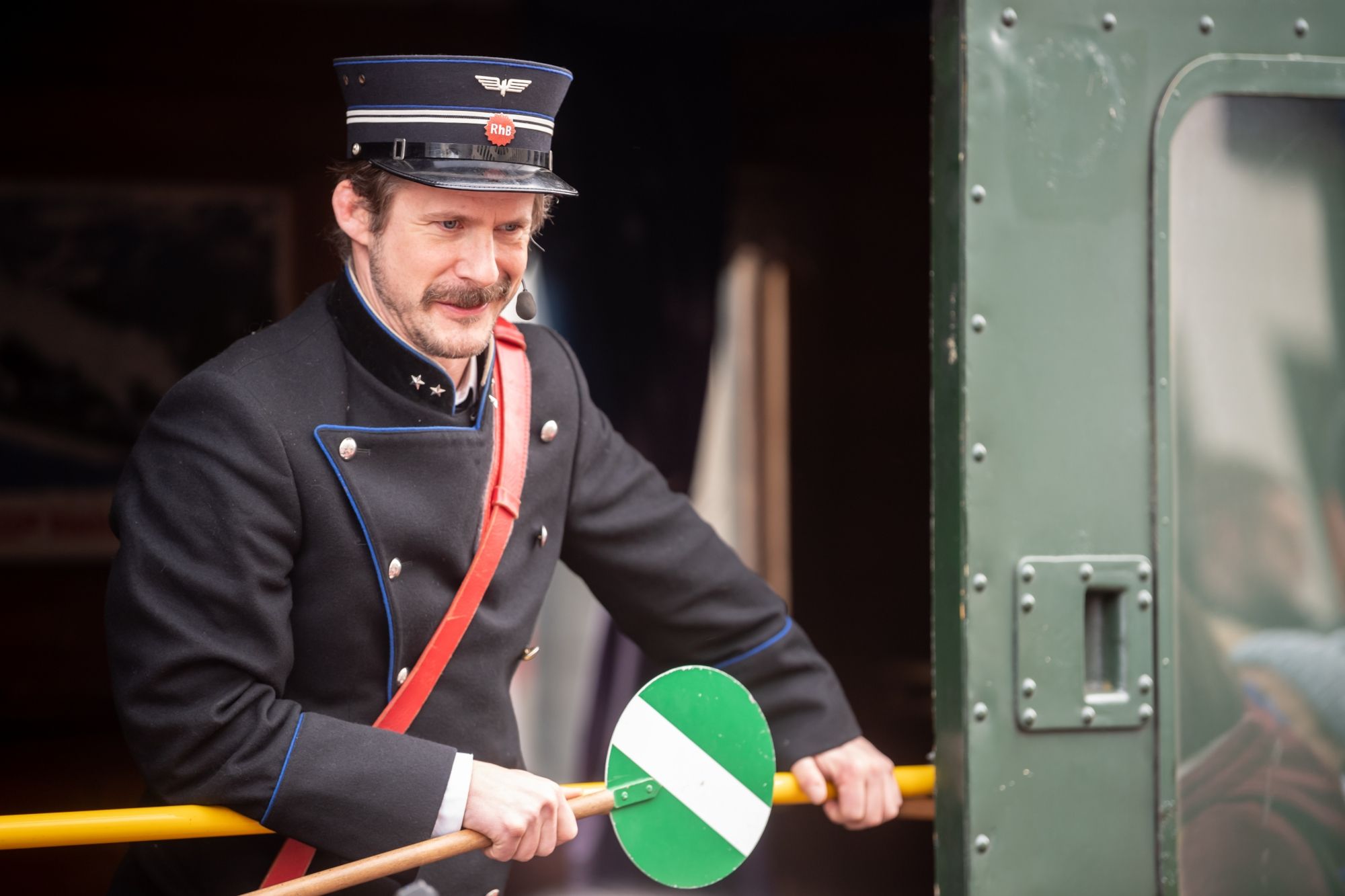 Linard Bardill en vêtements historiques, tenant un panneau de circulation vert, entouré de wagons de train
