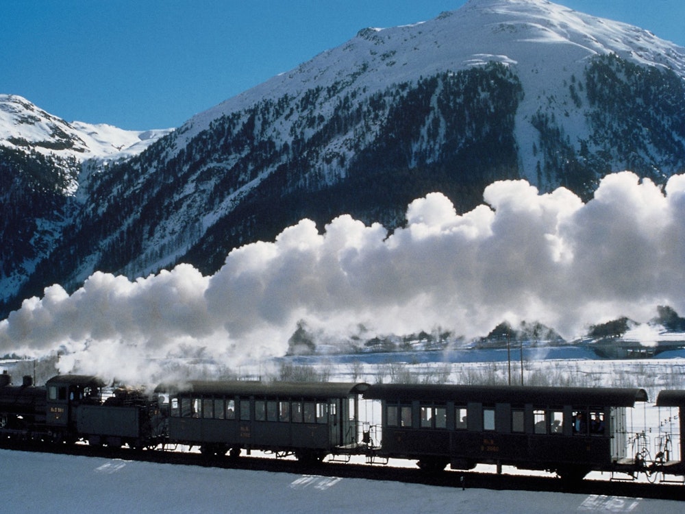 Engadin gőzmozdonyos utazás hóban, gőzmozdony és kocsik a hóesésben, havas hegyek előtt