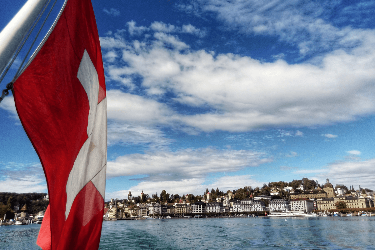 Charter do Dia: Vista do Lago de Lucerna com uma cidade pitoresca e a bandeira suíça.