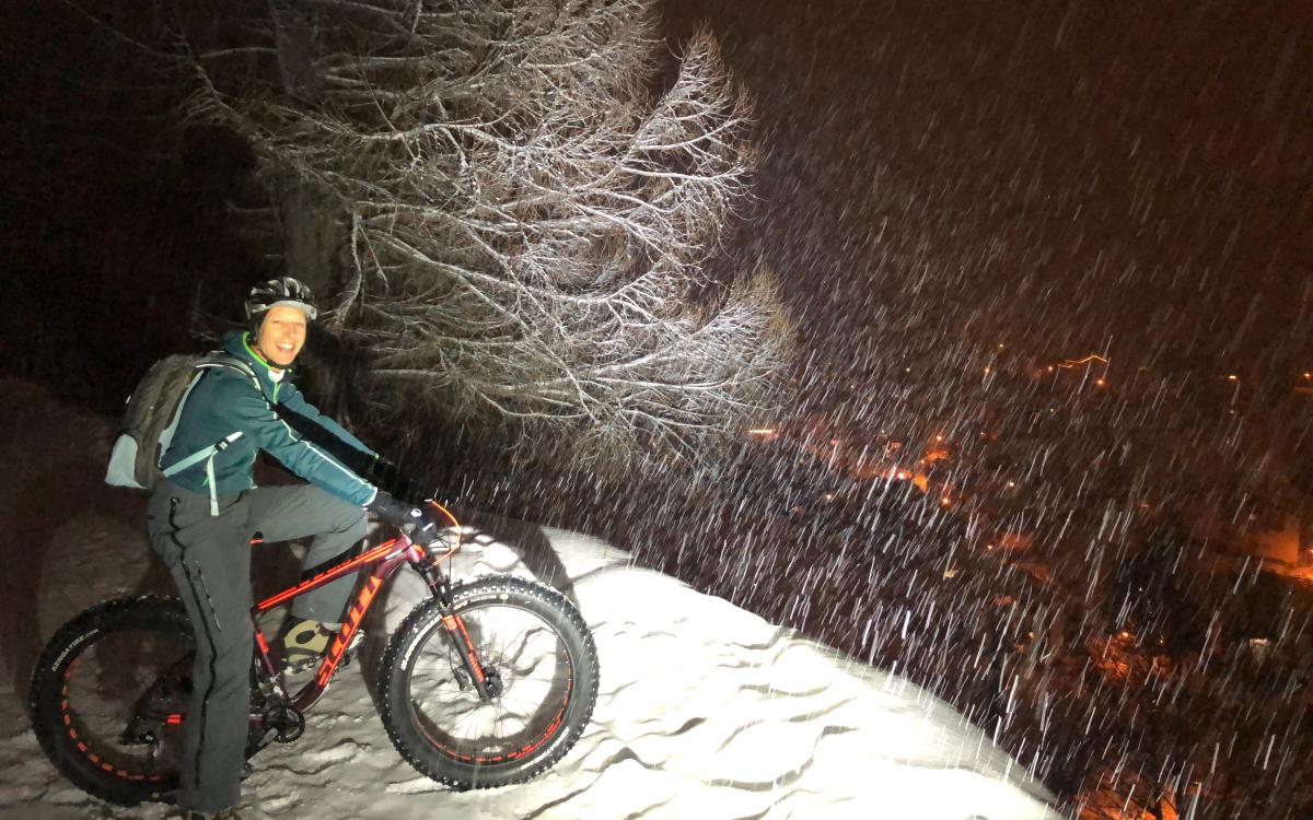 Kobieta jadąca na fatbike'u nocą po zaśnieżonych ścieżkach zimą, otoczona drzewami i śniegiem.