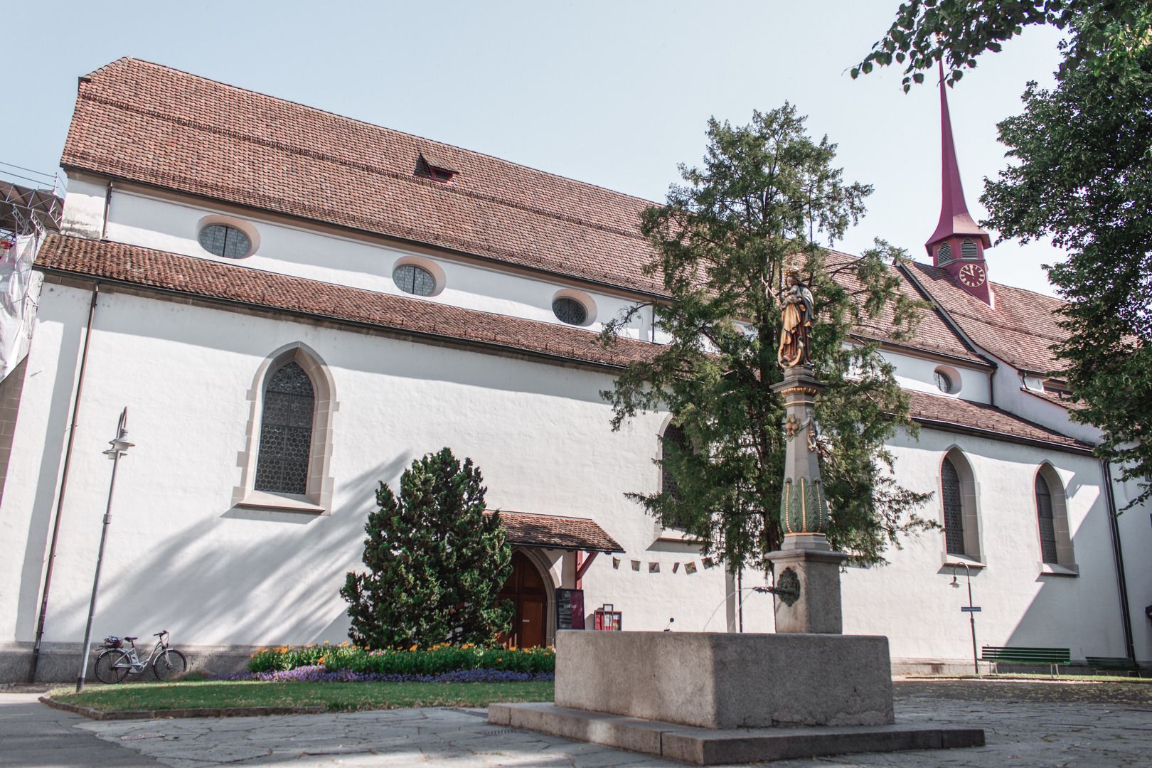 Franciskanerkyrkan i Luzern med en fontän i förgrunden. Besökare njuter av omgivningen i soligt väder.