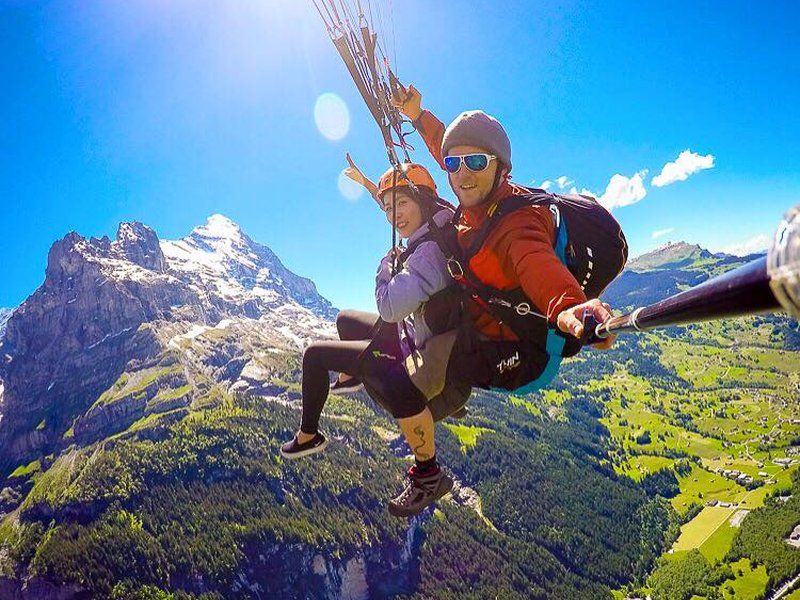 Parapente tándem en Grindelwald First con dos personas sobre los Alpes, visibilidad clara, cielo azul, prados verdes