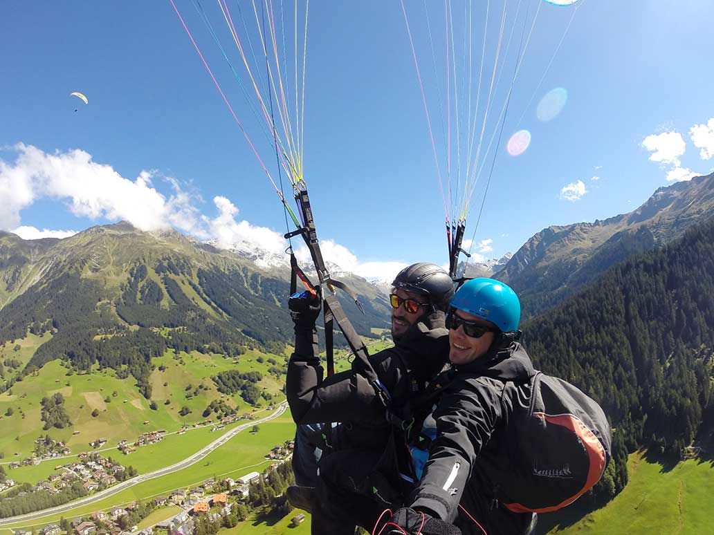 Paragliding ve dvojici v Davosu, dva lidé ve vzduchu, vidět horská krajina a zelená údolí.