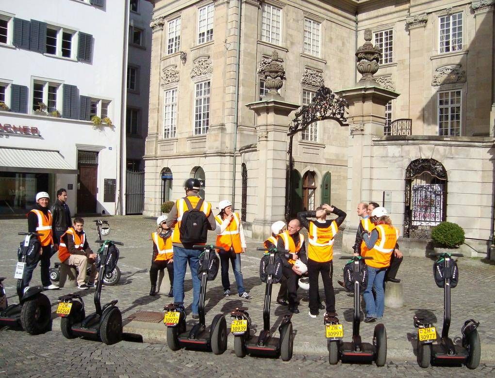 セグウェイツアー・チューリッヒは、歴史的な建物の前でオレンジ色のベストを着たグループを示しています。