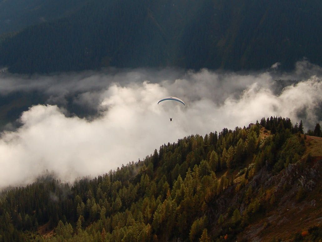 Paragliding ve dvojmístném letadle v Davosu, pilot plachtí nad mraky, zalesněné hory
