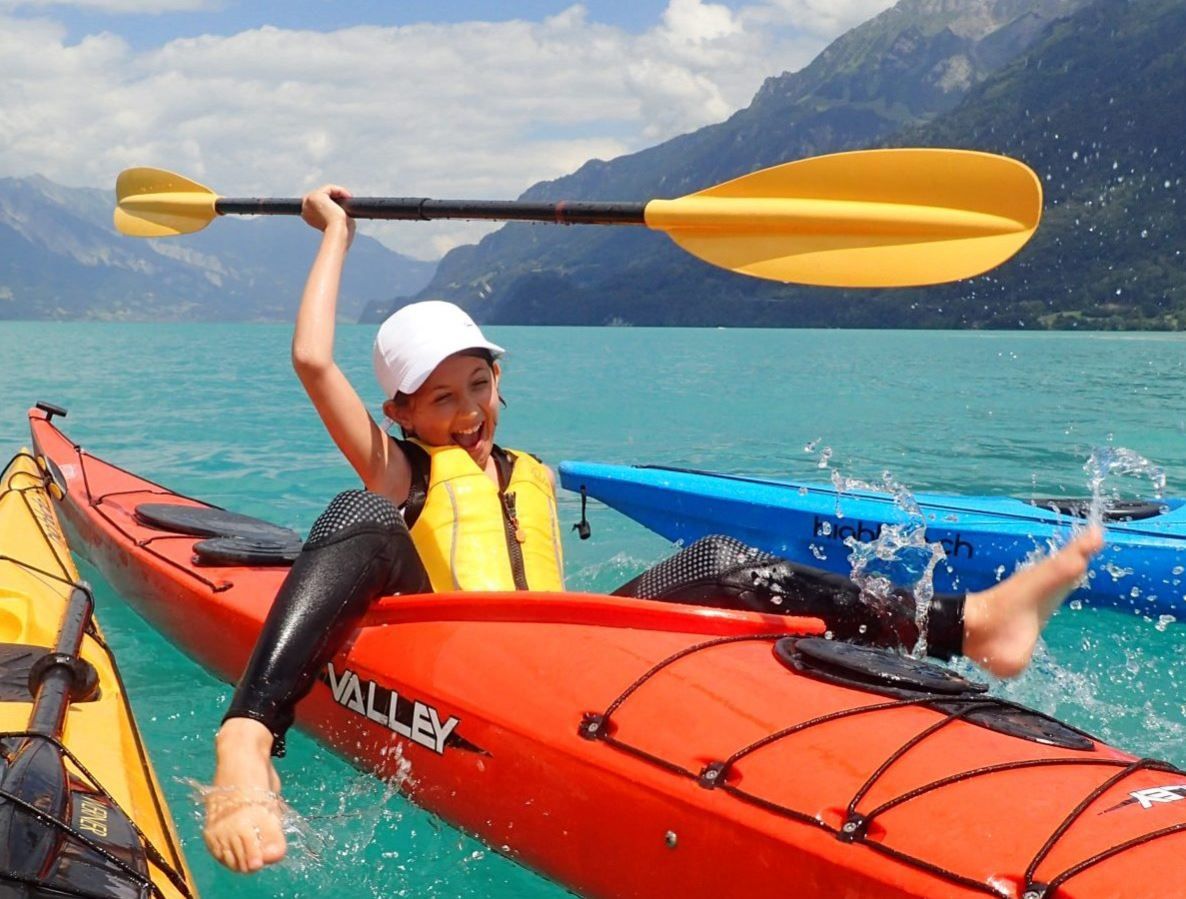 A child is paddling in a red kayak on a mountain lake and splashing water.