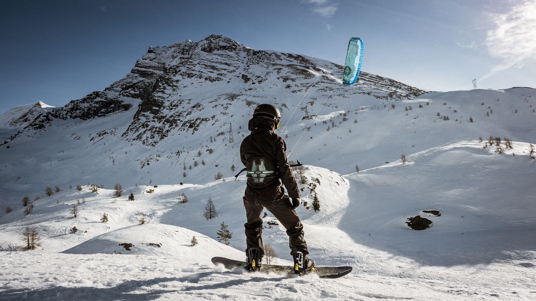 การเล่นสโนว์คิตในซิมพลอนสำหรับผู้เริ่มต้นพร้อมคนเล่นไคท์เซิร์ฟในหิมะ มีภูเขาอยู่เบื้องหลัง