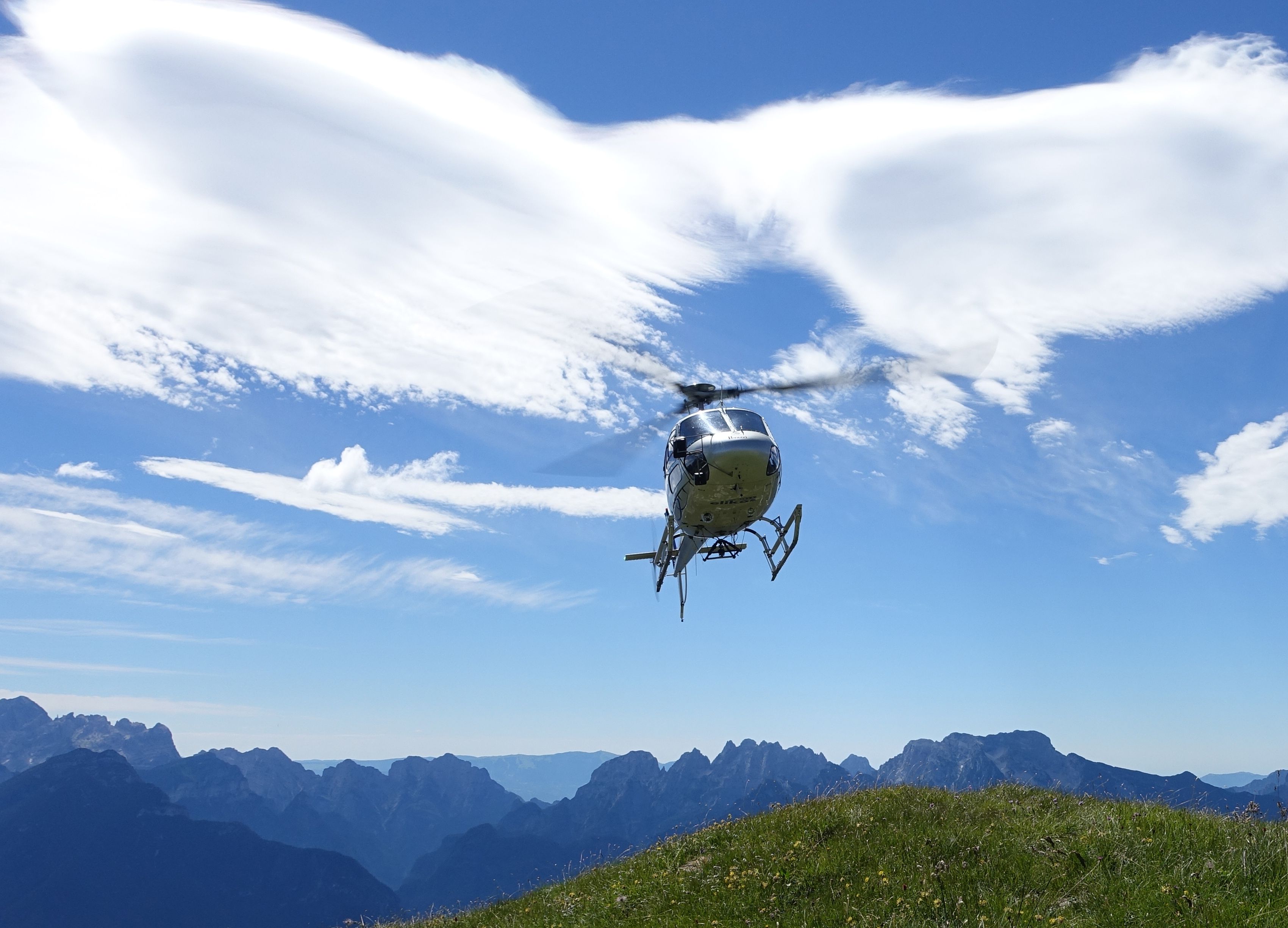 Helikopter Jura Seeland terbang di atas lanskap gunung di bawah langit biru.