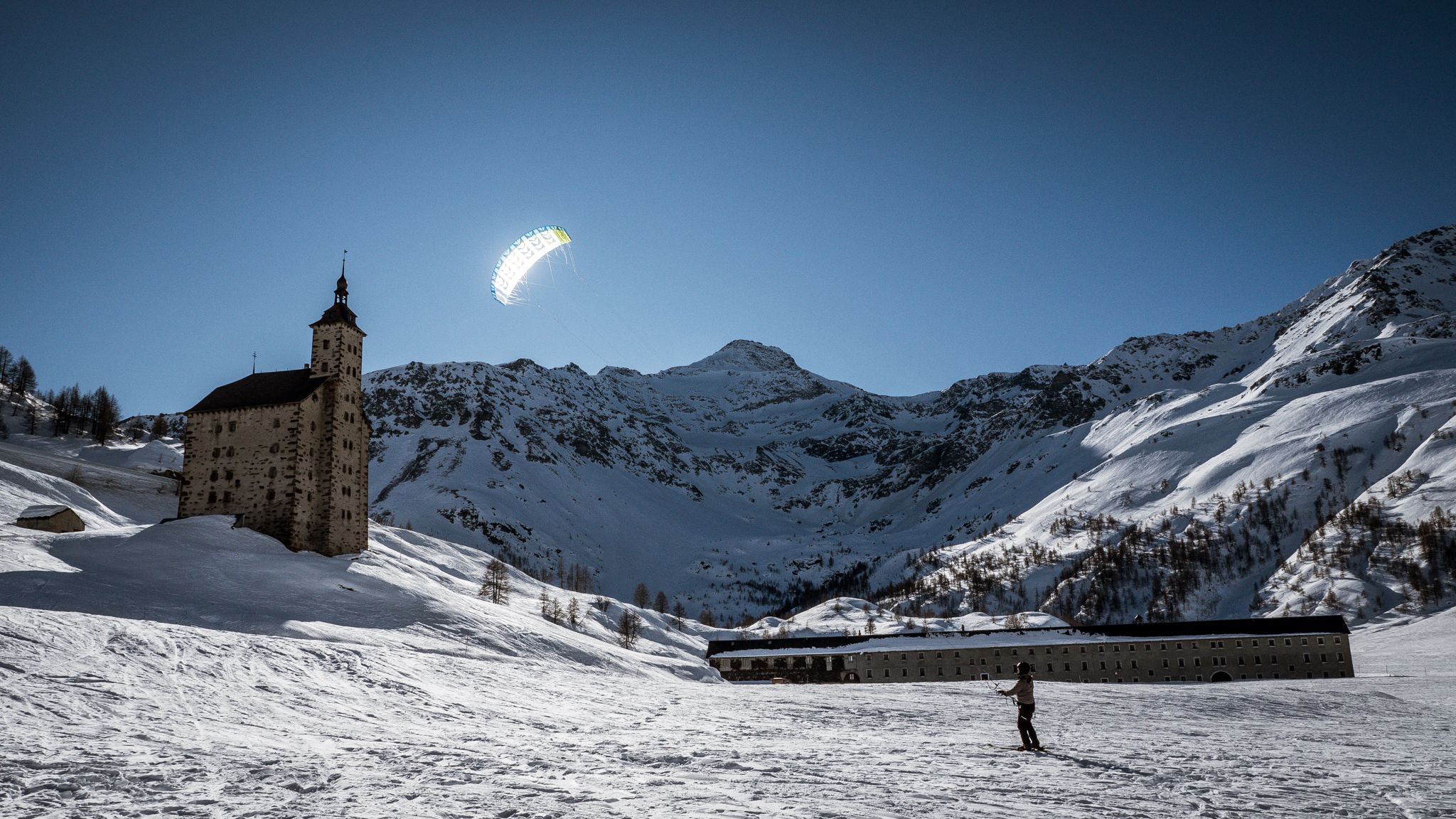 눈썰매 강습 심플론, 눈 덮인 들판 위에서 카이트 서핑하는 사람, 배경에는 건물과 산들이 있다.
