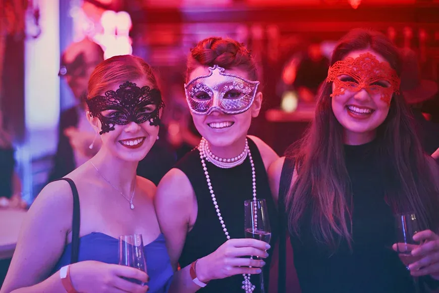 パーティー: マスクをつけた3人の女性がシャンパングラスを持ち、祝賀ムードの中で楽しんでいる。