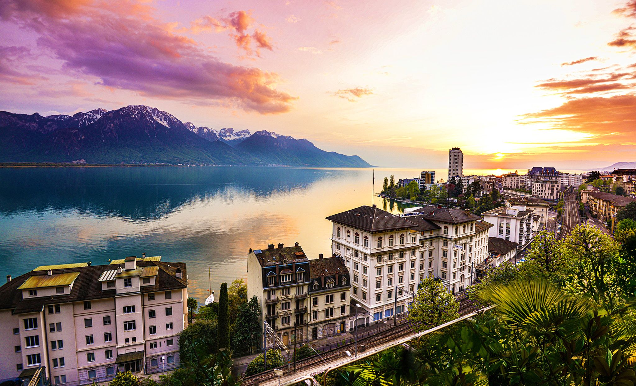 Montreux See mit Sonnenuntergang, reflektierende Wasseroberfläche, Gebäude und Berge im Hintergrund.
