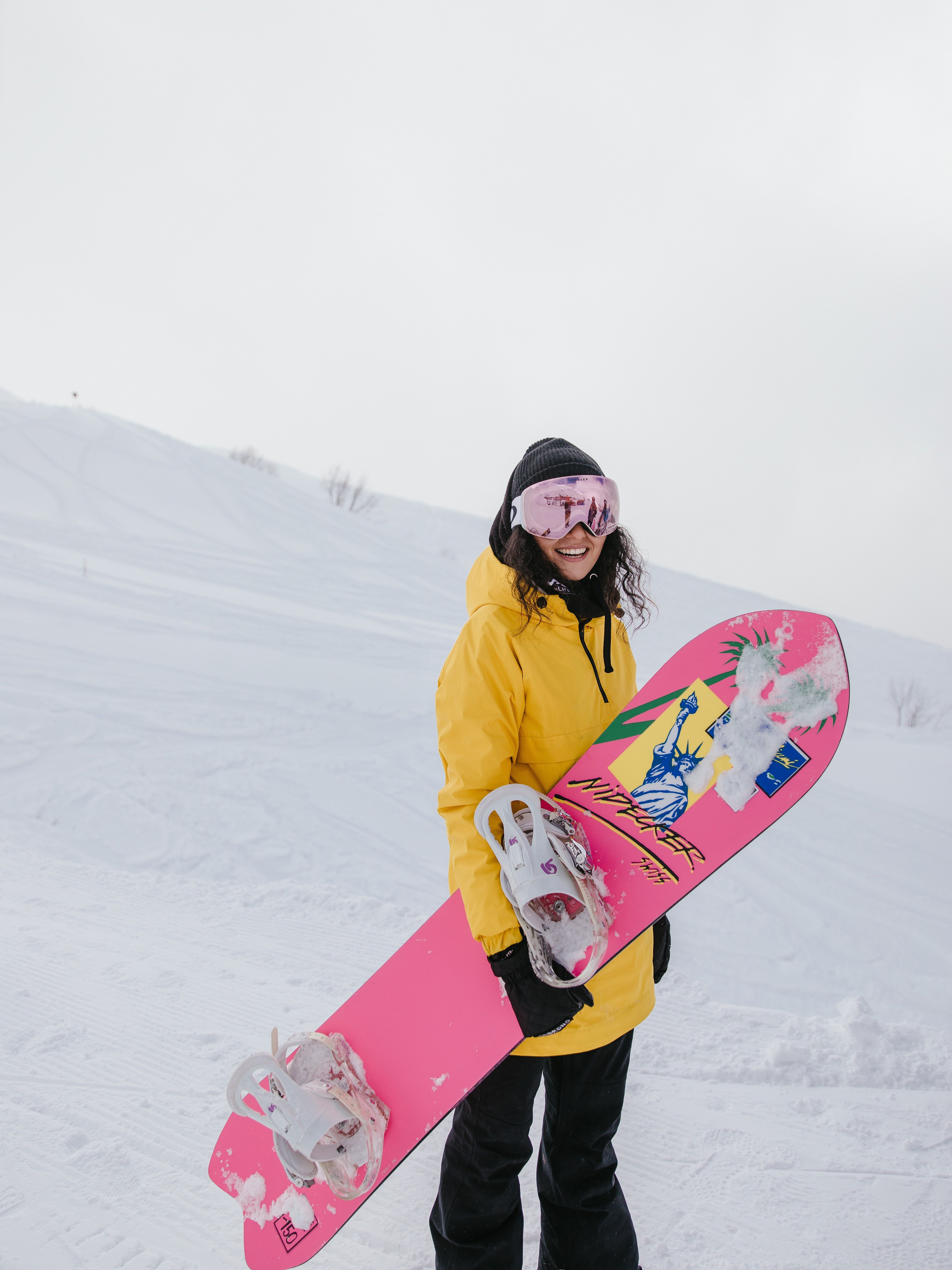 Snowboard: Wanita muda dengan pakaian kuning dan papan salju berwarna merah muda di medan bersalju.